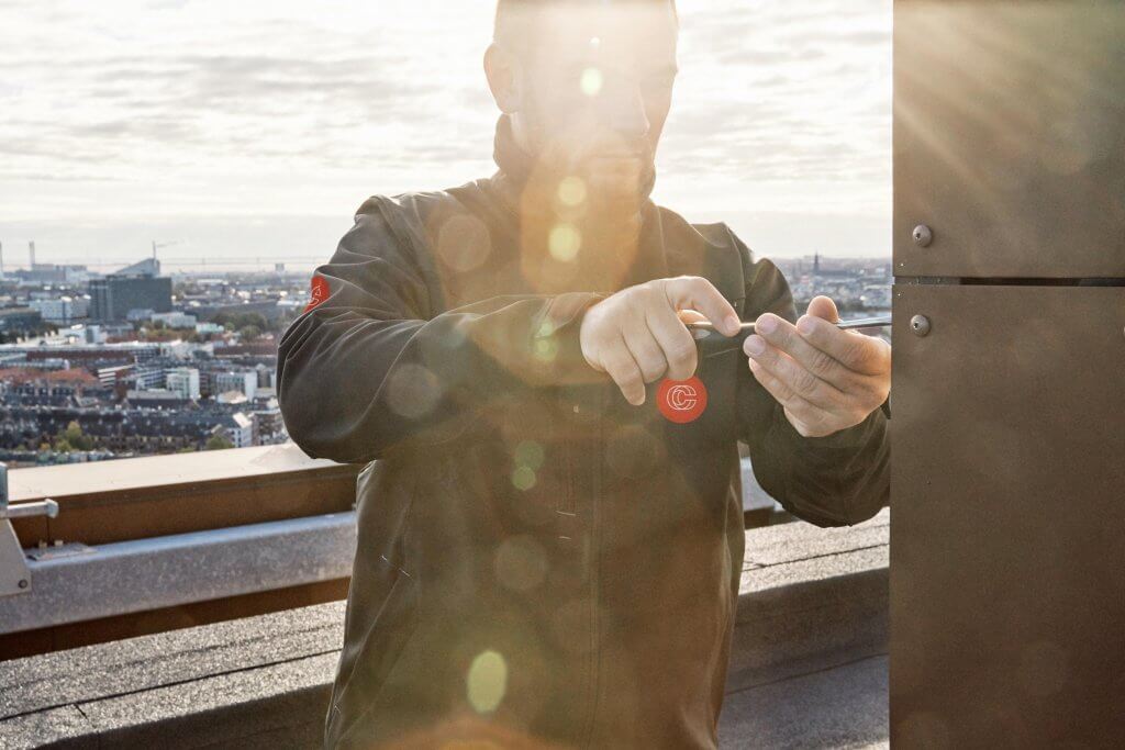 Mand med CC logo jakke på, der holder en skruetrækker i hånden og udfører ejendomsservice på taget af Nordbro, i København.